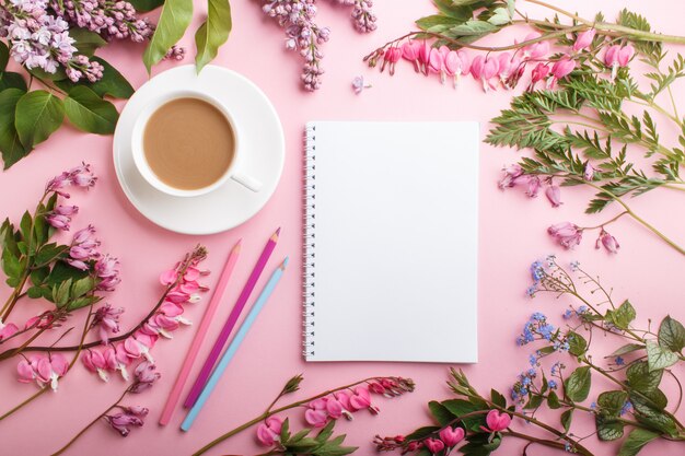 Lila lila und blutende herzblumen und ein tasse kaffee mit notizbuch und farbigen bleistiften auf pastellrosa.