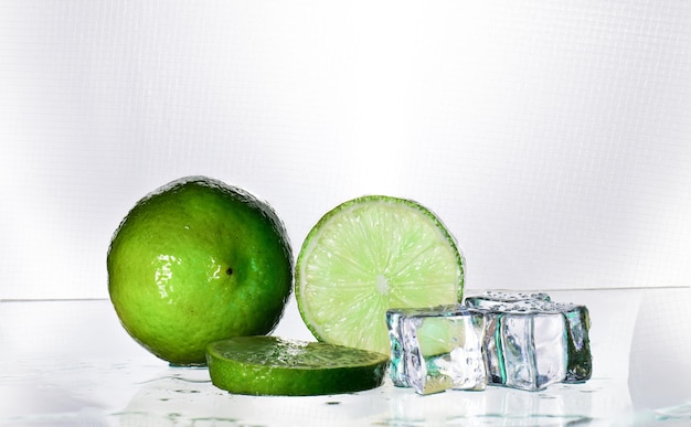 El limón verde está en blanco con una pieza cortada que muestra el interior de la ralladura de cítricos. El cristal transparente refleja las sombras del limón y el agua mojada dándole frescura y agregando hielo refrescante.