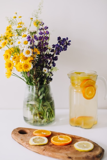 Limonada casera con naranja y limón y un ramo de flores naturales silvestres en mesa blanca