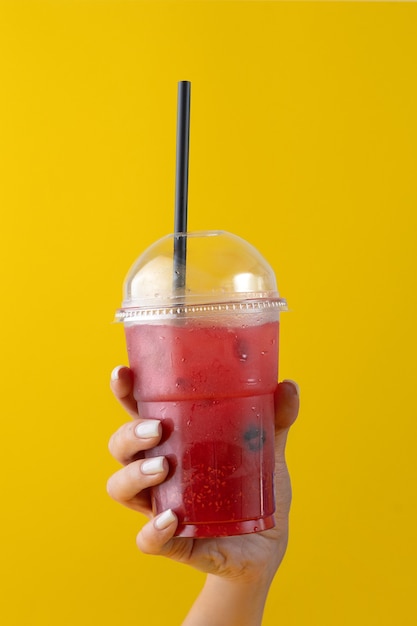 Limonada fresca de bebida de verão com morango no copo de plástico em fundo amarelo. mão feminina segurando um copo. Maquete para o seu anúncio.