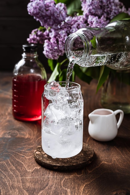 Limonada lilás refrescante em preparação Adicionando água com gás com gás Xarope lilás flores copo alto cheio de cubos de gelo