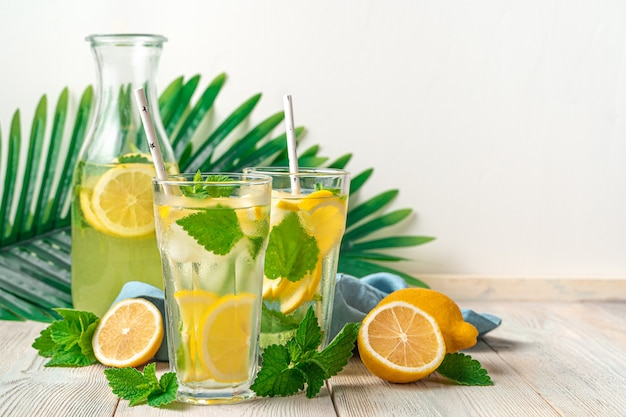 Limonada refrescante en dos vasos y un decantador, limones y menta sobre un fondo blanco con ramas de palmera. Vista lateral, copie el espacio.