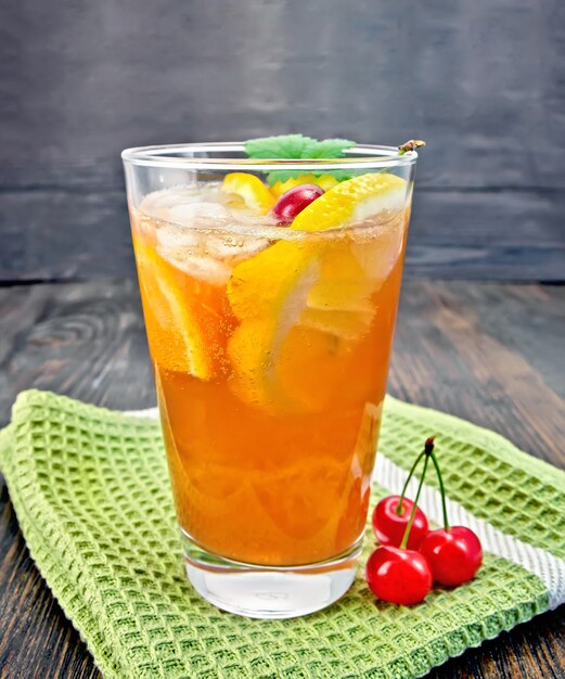 Limonada en un vaso con una cereza, limón y naranja, menta sobre una toalla verde sobre un fondo de una tabla de madera oscura.