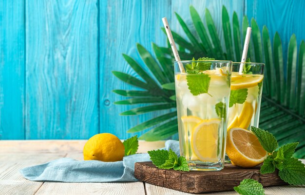 Limonade mit Eis, Minze und Zitronenscheiben in gedämpften Gläsern auf einem Hintergrund von Palmblättern. Seitenansicht, Kopienraum.