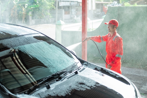 Limpador de carro masculino usando uniforme vermelho borrifa água no carro enquanto lava o carro