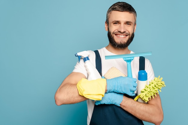 Limpador feliz em uniforme e luvas de borracha segurando material de limpeza em fundo azul