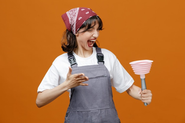 Limpador feminino jovem irritado vestindo uniforme e bandana segurando o êmbolo olhando para ele mantendo a mão no ar gritando isolado em fundo laranja