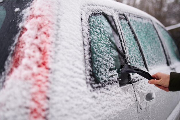 Limpando as janelas laterais do carro de neve com raspador de gelo antes da viagem Homem remove gelo das janelas do carro Mão masculina limpa o carro com ferramenta especial no dia de inverno gelado de neve