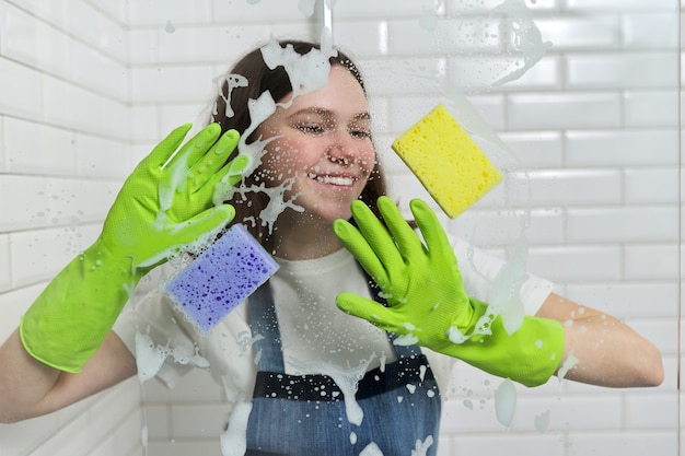 Limpando o banheiro, menina adolescente lavando o vidro do chuveiro com espuma e esponjas, close-up de mulheres com luvas verdes e esponjas coloridas