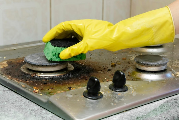 Limpar o fogão a gás sujo das sobras de comida gordurosa deposita a mão da mulher na luva protetora com pano de esponja e detergente lavando o conceito de serviço de limpeza doméstica do fogão de cozinha