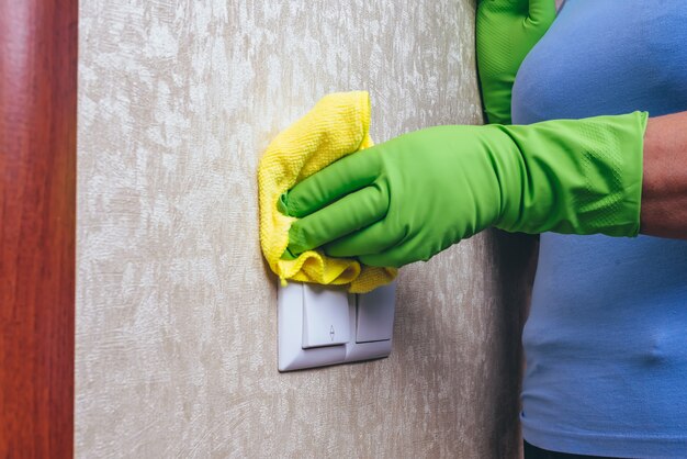 Limpeza em casa. Uma garota de luvas verdes limpa o interruptor de eletricidade com um pano amarelo