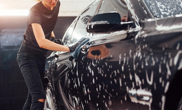 Limpia el vehículo que está en jabón blanco. Una mujer limpia un automóvil negro moderno dentro de la estación de lavado de autos.