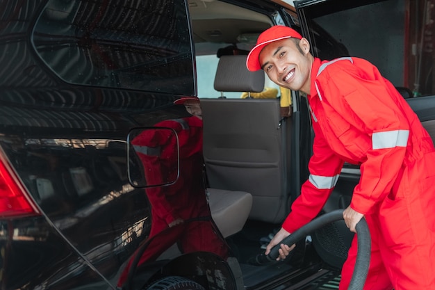 Limpiador de automóviles masculino asiático viste uniforme rojo sonriente mientras limpia el piso del automóvil en el salón del automóvil