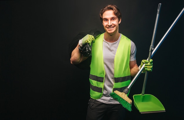 El limpiador europeo sonriente sostiene la bolsa de basura, la escoba y la pala. Hombre guapo joven usar uniforme verde y guantes de goma. Concepto de servicio de limpieza. Aislado sobre fondo oscuro. Sesión de estudio. Copia espacio