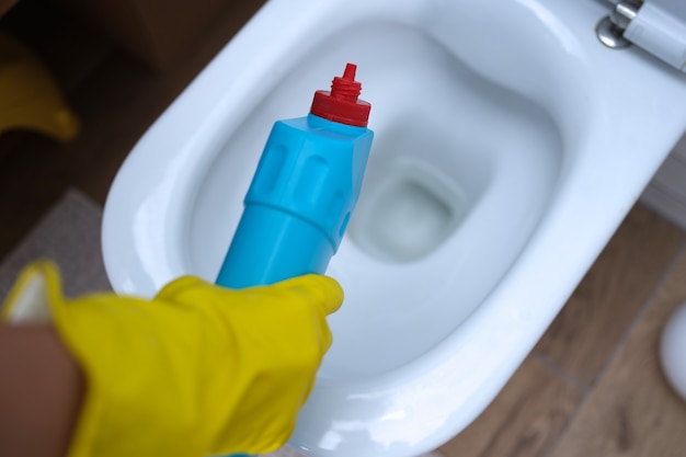 Limpiador en guantes de goma amarillos vertiendo detergente debajo del borde del inodoro closeup