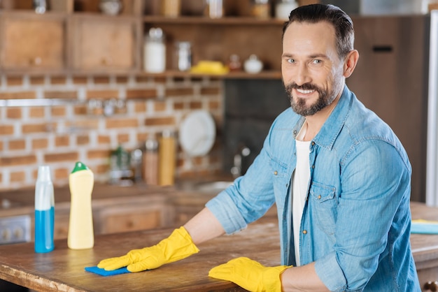 Limpiador masculino vigoroso positivo mirando directamente mientras sonríe y frota el escritorio