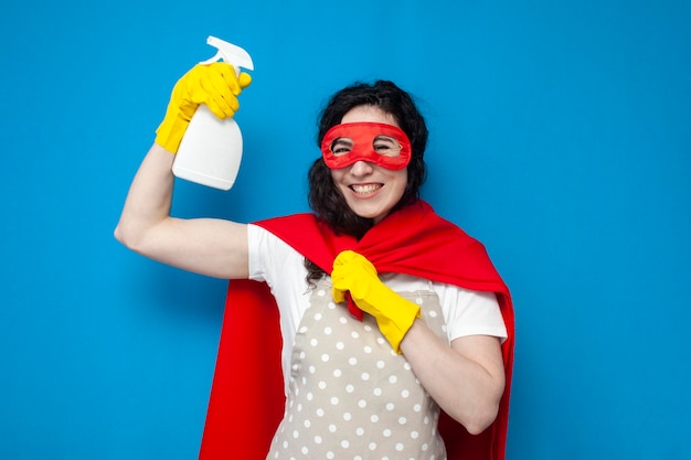 Limpiador de niña en guantes y traje de superman tiene detergente sobre fondo azul.