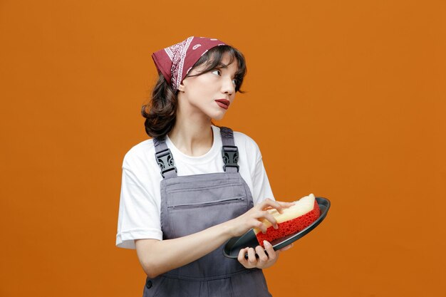 Limpiadora joven impresionada con uniforme y bandeja de limpieza de bandana con esponja mirando al lado aislado en fondo naranja