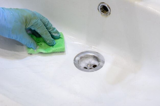 Limpiando el baño. La mujer está limpiando el fregadero y el grifo con una esponja en guantes de goma y detergente en aerosol. De cerca. Copie el espacio.