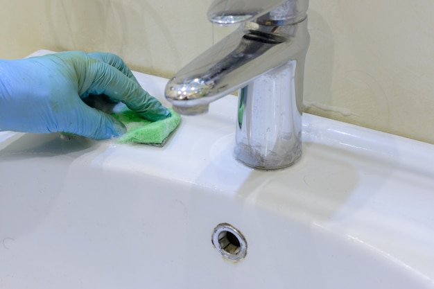 Limpiando el baño. La mujer está limpiando el fregadero y el grifo con una esponja en guantes de goma y detergente en aerosol. De cerca. Copie el espacio.