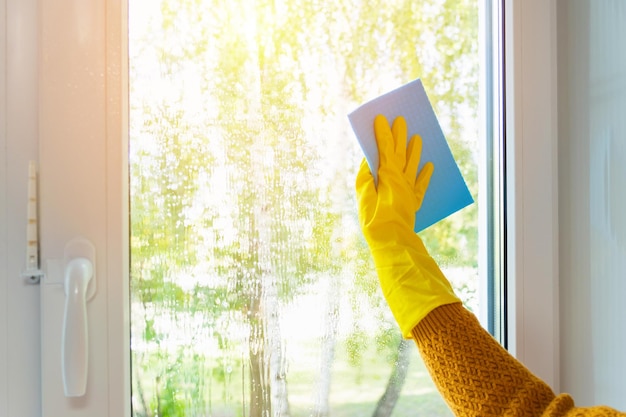 Limpiando la casa. La mano de una mujer con guantes de goma amarillos lava la ventana con un trapo azul.