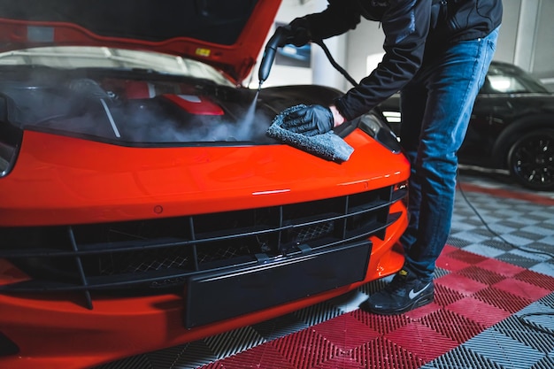 Limpiar un coche moderno de color naranja con vapor y paño de microfibra en el taller de reparación de automóviles