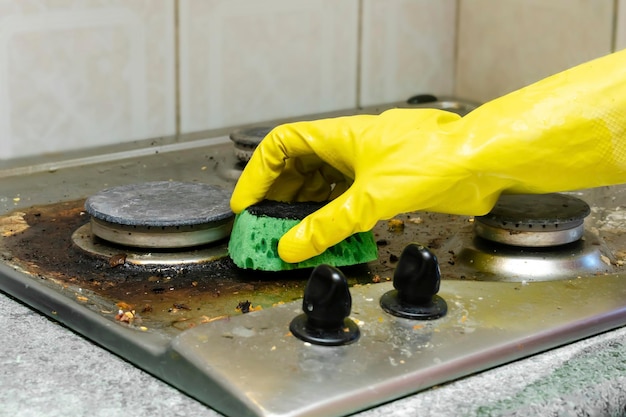 Limpiar la estufa de gas sucia de grasa, restos de comida y depósitos. Mano de mujer en guante protector con trapo de esponja y estufa de cocina de lavado con detergente. concepto de servicio de limpieza a domicilio.