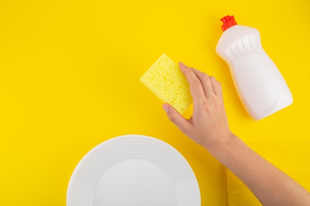 Limpieza de esponja amarilla sosteniendo la mano y detergente para platos sobre fondo amarillo con placas blancas