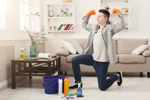 Limpieza de primavera hecha. Hombre feliz con las manos levantadas de pie sobre una rodilla en apartamento limpio entre detergentes, balde y plumeros. Hogar y concepto de limpieza.