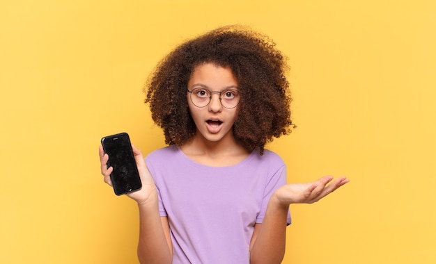 Linda adolescente afro se sentindo extremamente chocada e surpresa, ansiosa e em pânico, com um olhar estressado e horrorizado e segurando um celular