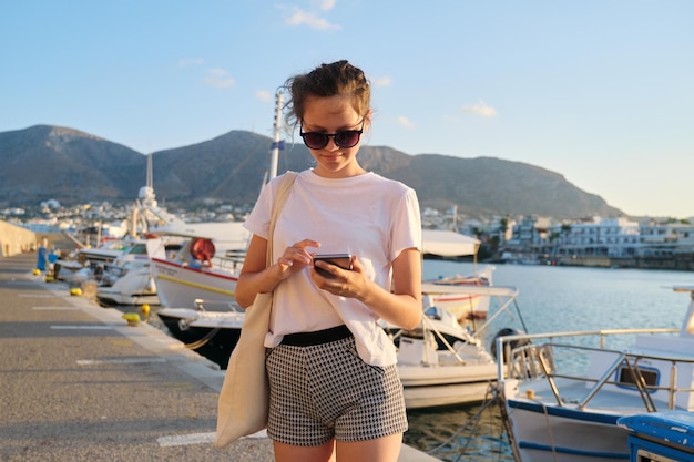 Linda adolescente caminhando ao longo do cais com iates ancorados, adolescente com smartphone, fundo de paisagem de montanha do pôr do sol do mar, férias de verão, viagem no mar