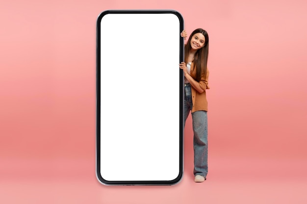 Linda adolescente espiando atrás de um enorme smartphone em branco com tela branca
