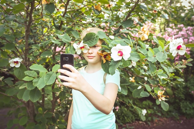 Linda adolescente loira faz selfie no fundo das árvores de magnólia da primavera florescendo com flores brancas perfumadas