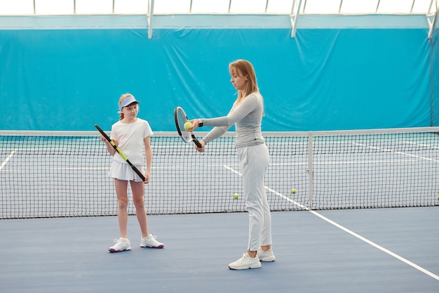 Linda adolescente en ropa deportiva mirando a la entrenadora y escuchando sus instrucciones sobre cómo usar la pelota y la raqueta durante el juego