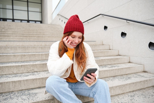 Linda adolescente ruiva senta-se na escada e sorri segura telefone celular usa aplicativo de smartphone