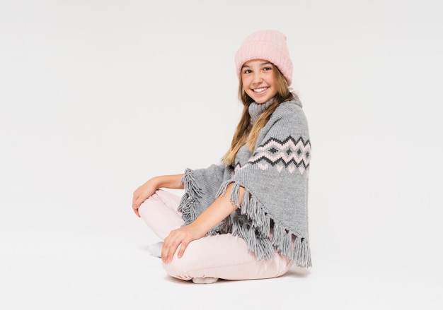 Linda adolescente sonriente con gorro de punto rosa y poncho gris acogedor aislado en el fondo blanco