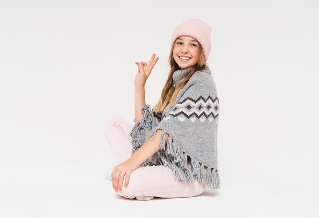 Linda adolescente sonriente con gorro de punto rosa y poncho gris acogedor aislado en el fondo blanco