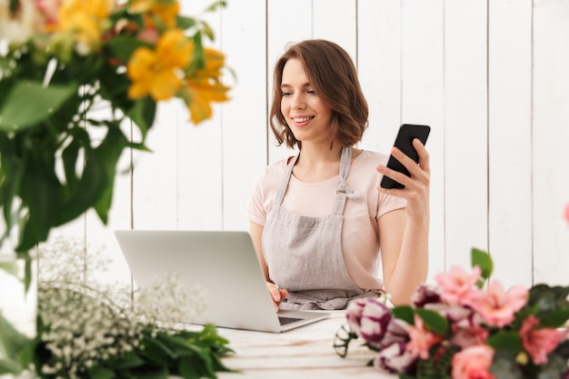 Linda alegre florista dama usando la computadora portátil chateando por teléfono móvil.