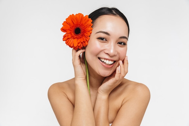 linda alegre jovem muito asiática com pele saudável posando nua isolada sobre uma parede branca, segurando uma flor.