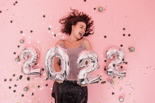 Linda alegre menina morena vestida festivamente com cabelo encaracolado em um fundo rosa com balões de prata dos números 2022, conceito de festa de ano novo.