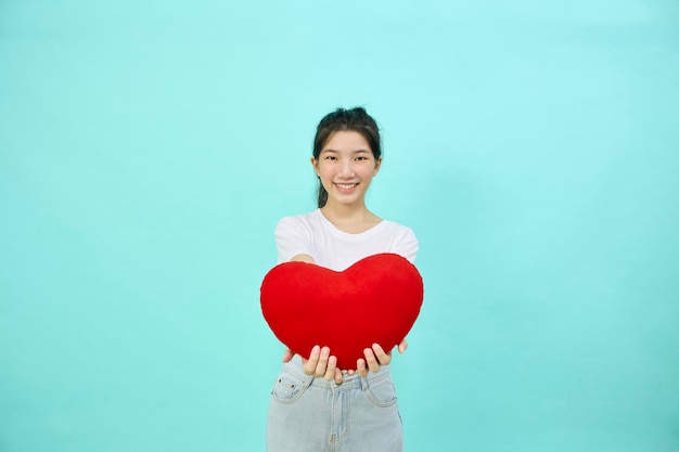 Linda alegre mujer asiática de pelo negro sonríe con forma de almohada de corazón sobre fondo de estudio azul claro