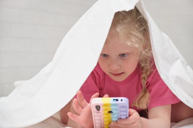 Una linda y alegre niña en edad preescolar yace debajo de la manta con un teléfono inteligente en las manos