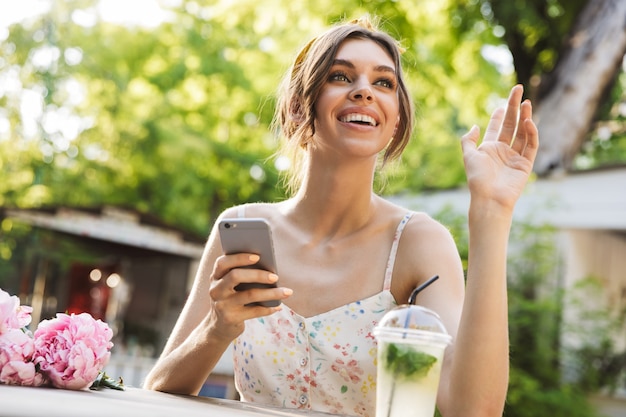 linda alegre sorridente jovem incrível mulher sentada à mesa com flores no parque verde, usando telefone celular, acenando para um amigo.