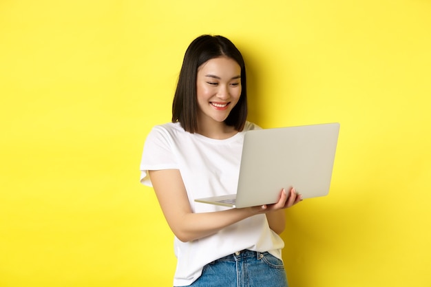 Linda aluna asiática trabalhando no laptop, lendo a tela e sorrindo, fundo amarelo.