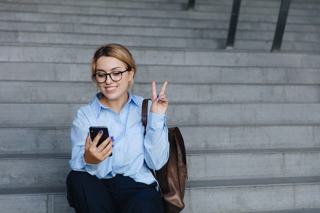 Linda aluna com cabelo loiro, mostrando sinal de paz com os dedos ao tirar selfie no smartphone. Conceito de tecnologia moderna.