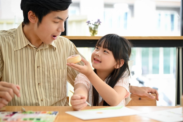 Una linda y amable niña asiática comparte un donut con su papá en el café feliz tiempo familiar