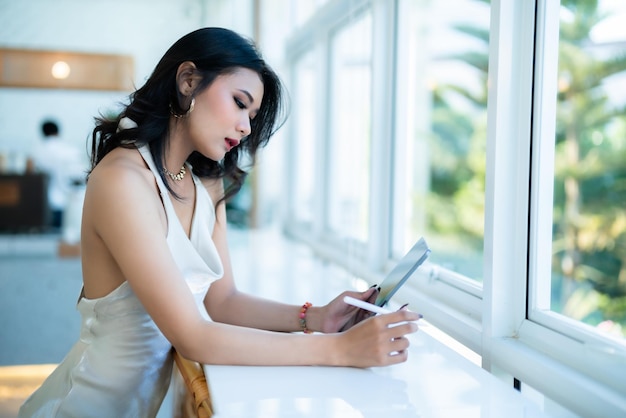 Linda asiática freelance pessoas sucesso modelo de moda mulher de negócios no verão vestido branco sem mangas usando casual trabalhando com tablet para navegar na internet conversando e blogando no café