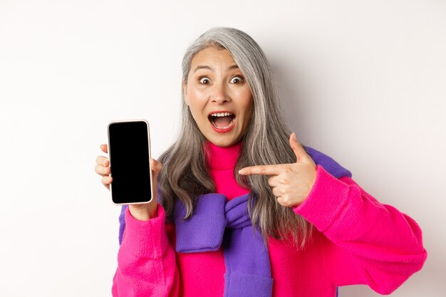 Linda avó asiática sorrindo, apontando o dedo para a tela em branco do smartphone, parecendo espantada, mostrando o aplicativo móvel, em pé no branco.