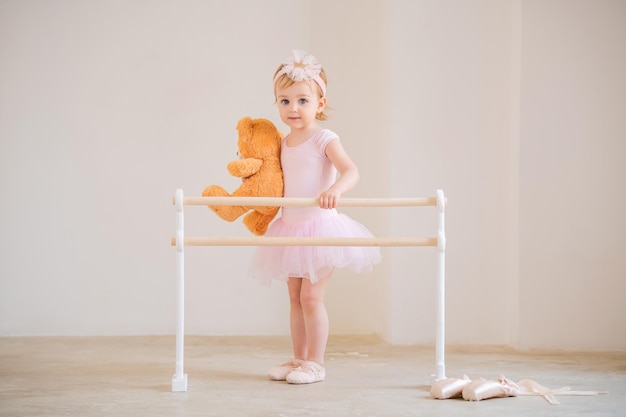 Linda bailarina de olhos azuis em pé rosa perto da barra de balé com um ursinho de pelúcia nas mãos Conceito de sonho de se tornar uma bailarina