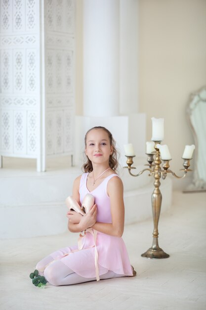 Una linda bailarina en un traje de ballet rosa está sentada en el suelo con punta en sus manos y sonriendo. Chica en la clase de baile. La niña está estudiando ballet. Ballet clásico, danza. Estudio de ballet.